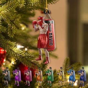 May include: A Christmas ornament featuring a boxer in red shorts, gloves, and headgear, posed next to a red punching bag with the name "DAVID". The ornament is hanging from a gold string, with several similar ornaments in different colors below.
