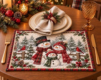 May include: A rectangular quilted placemat featuring a printed winter scene of three snowmen wearing hats and scarves, surrounded by pine trees, red cardinals, and holly berries. The placemat has a red and green plaid border and a stitched texture.