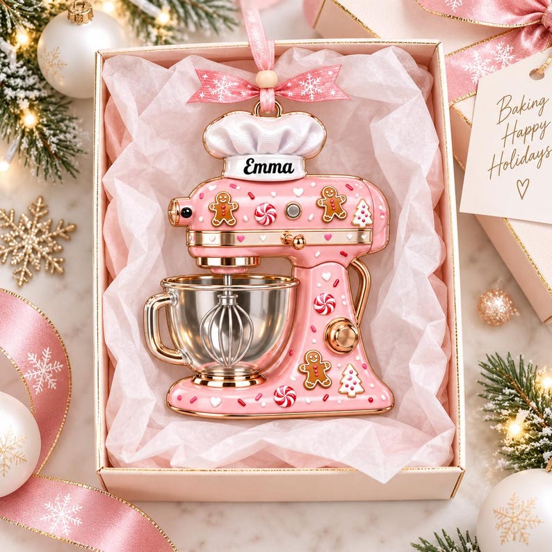 A pink and gold Christmas ornament in a box, featuring a silver mixer and gingerbread men, surrounded by holiday decorations such as snowflakes and ornaments.