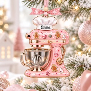 A pink and gold Christmas ornament shaped like a mixer, with gingerbread men and other festive decorations, hanging on a tree.