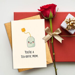 May include: A greeting card with a cute tea bag illustration and the text "You're a TEA-RIFFIC MOM." The card is next to a red rose with a gold ribbon, a red gift box, and a small white gift box with a gold bow.