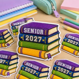 May include: Embroidered patches in the shape of stacked books, featuring the text "SENIOR 2027" in purple, blue, green, and yellow. The patches have a gold border and a small purple bookmark. Other school supplies are in the background.