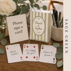 May include: A clear plastic box containing a deck of cards with sage green stripes and an oval label reading Daniel & Sophia Est. 2024. Beside it, a sign reads Please sign a card! and three playing cards display the text Best wishes!, So happy for you!, and Cheers to forever!.