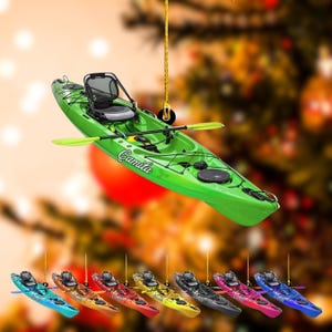 May include: A green kayak ornament with a black seat and paddle. The kayak has the name "Camila" printed on the side. Below, there are several smaller kayak ornaments in various colors, including blue, orange, red, yellow, black, pink, and blue.