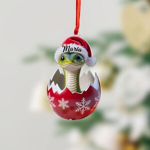 May include: A Christmas ornament featuring a green snake emerging from a red eggshell decorated with snowflakes. The snake wears a Santa hat with the name "Maria" on it, suspended by a red ribbon.