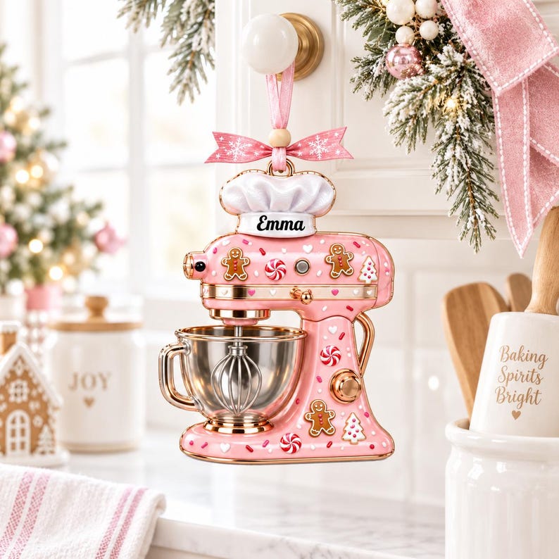 A pink and gold kitchen ornament featuring a mixer and gingerbread man decorations, with a pink ribbon and the name "Emma" on the front.
