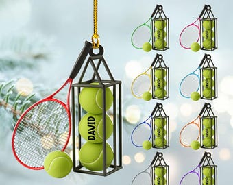 May include: A tennis-themed ornament featuring a black cage holding three green tennis balls with the name "DAVID" printed on them. A red tennis racket hangs next to the cage. Additional ornaments with different colored rackets are also shown.