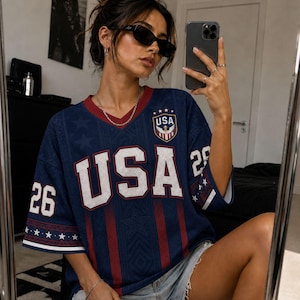 A woman wearing a blue USA jersey with the number 26 is taking a selfie in front of a mirror.