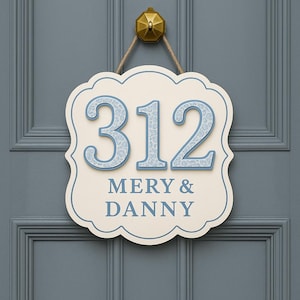 May include: A cream-colored house number sign with a scalloped edge, displaying the number "312" in light blue with a leopard print pattern. The names "MERY & DANNY" are below the numbers. The sign hangs from a gold-colored knob.