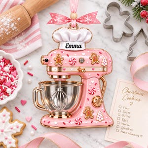 a pink and gold Christmas ornament shaped like a mixer, decorated with gingerbread men, candy canes, and other festive elements. The ornament is surrounded by various baking-related items, such as a cookie cutter, a bowl of sprinkles, and a note with a list of ingredients for making Christmas cookies.