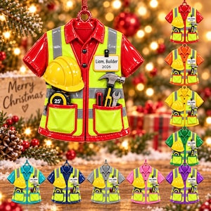 May include: A red construction shirt ornament featuring a yellow hard hat, a 25' tape measure, a hammer, and a wrench. A white name tag on the chest reads Liam, Builder 2026. The shirt has reflective silver stripes and two pockets.