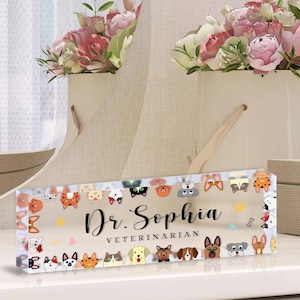 May include: A clear acrylic desk nameplate for a veterinarian, featuring the name "Dr. Sophia" and the word "VETERINARIAN". The design includes a border of cartoon dog faces in various colors and styles.
