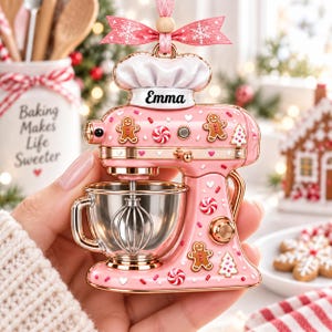 A hand holding a pink and gold ornament shaped like a pink and gold mixer with gingerbread men and other festive decorations.
