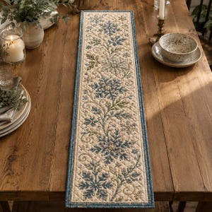 May include: A rectangular quilted table runner featuring a cream-colored background with a repeating pattern of blue and green embroidered snowflakes and swirling vine motifs, finished with a solid blue border and a diamond-stitched texture.