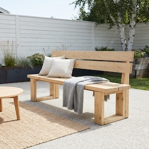 May include: A light-colored wooden bench with a backrest and rectangular legs. Two off-white square throw pillows are placed on the seat, and a soft gray throw blanket is draped over the edge of the bench, partially covering the wooden surface.