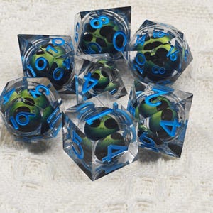 May include: Set of seven clear, blue-rimmed dice with green and black alien faces. The dice are numbered 1 through 6.