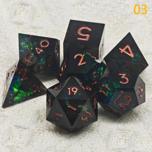 Puede incluir: Conjunto de siete dados iridiscentes negros y verdes con n&uacute;meros de cobre. Los dados son un d4, d6, d8, d10, d%, d12 y d20.