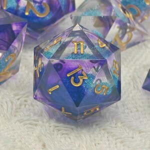 May include: A set of polyhedral dice with a clear resin exterior, showcasing a blend of blue, purple, and teal colors. The dice feature gold-colored numbers and are designed for tabletop gaming.