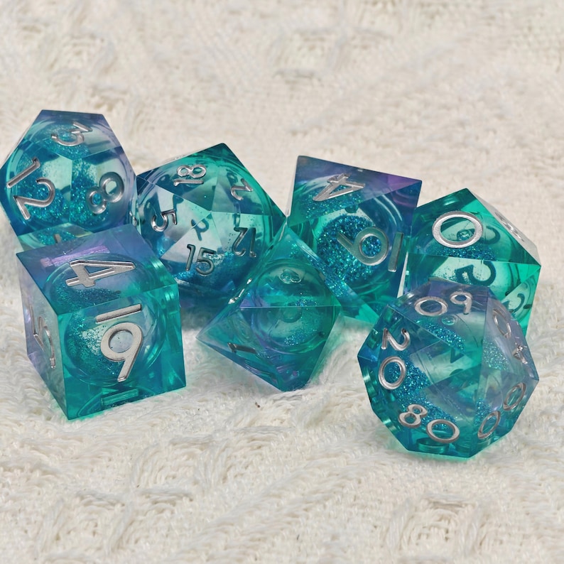 Puede incluir: Conjunto de siete dados iridiscentes azul y verde con n&uacute;meros plateados. Los dados son transl&uacute;cidos y tienen un acabado brillante.