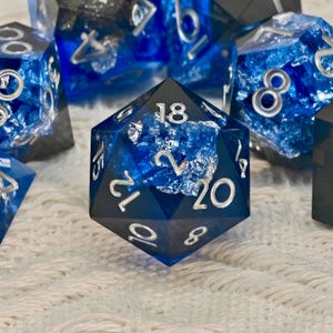 Puede incluir: Primer plano de un conjunto de dados poliédricos. Los dados son de color azul oscuro con números plateados e inclusiones internas. Los dados están colocados sobre una superficie blanca y texturizada.