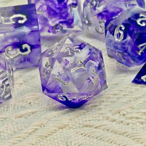 Puede incluir: Un conjunto de dados poliédricos con un diseño veteado translúcido púrpura y blanco. Los dados tienen numeración plateada y están diseñados para juegos de rol. El conjunto incluye varias formas, incluido un dado de veinte caras.