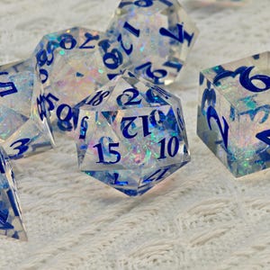May include: A collection of clear, multi-sided dice with blue numbering and iridescent glitter inclusions. The dice are scattered on a white, textured surface, showcasing their geometric shapes and reflective qualities. Ideal for tabletop gaming.