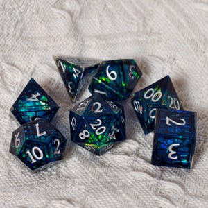 May include: A set of seven polyhedral dice featuring a dark blue and green translucent resin base with embedded iridescent foil shards and crisp white numerals. The collection includes a d20, d12, d10, d8, d6, d4, and a percentile die.