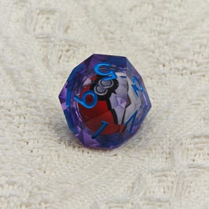 May include: A purple and blue 12-sided die with a Pokeball design inside. The die has blue numbers on each side.