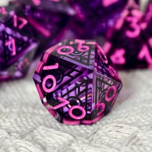 Könnte beinhalten: Ein zwanzigseitiger Würfel aus durchscheinendem violettem Harz mit einem eingebetteten schwarzen geometrischen Muster. Die Zahlen sind eingraviert und mit leuchtend neonpinkem Pigment gefüllt, das sich vom dunkelvioletten und schwarzen Design abhebt.