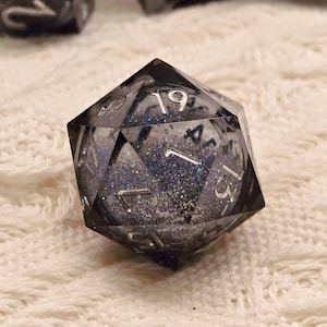 Puede incluir: Un dado transparente de múltiples caras con números plateados y un interior oscuro y brillante. El dado es de color gris oscuro y está sobre una superficie blanca texturizada. Los números son claramente visibles, para juegos de mesa.