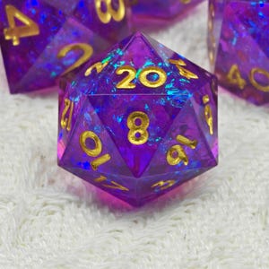 May include: A purple iridescent 20-sided die with gold numbers. The die is made of a clear material with a glittery finish.