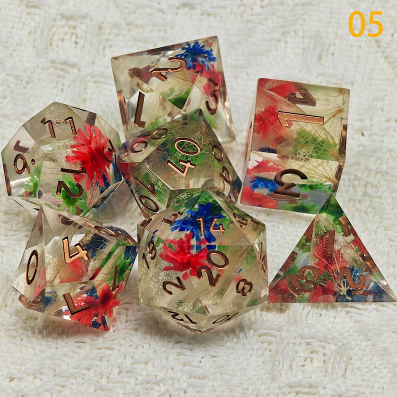 Puede incluir: Conjunto de seis dados de resina transparente con flores incrustadas y n&uacute;meros dorados. Los dados son de varias formas, incluyendo un d4, d6, d8, d10, d12 y d20.