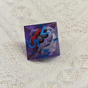 May include: A purple, clear, six-sided dice with blue numbers. The dice has a white center with a black and red design.