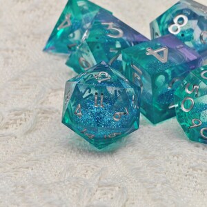 Puede incluir: Un conjunto de seis dados de color azul turquesa con n&uacute;meros plateados. Los dados est&aacute;n hechos de pl&aacute;stico transparente con purpurina incrustada en el pl&aacute;stico. Los dados est&aacute;n sobre una superficie blanca y peluda.