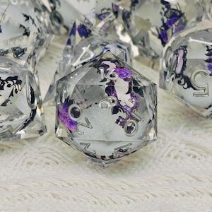May include: Set of clear, multi-sided dice with silver numbering and purple floral designs. The dice are filled with a glittery substance, creating a shimmering effect. The dice are for tabletop games.