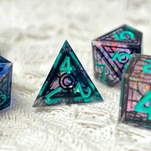 May include: A set of translucent polyhedral dice featuring a dark, smoky resin base embedded with iridescent, shimmering flakes. Each die has bold, bright teal numbers and intricate black geometric line work etched into the surface.