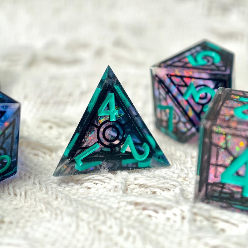 May include: A set of translucent polyhedral dice featuring a dark, smoky resin base embedded with iridescent, shimmering flakes. Each die has bold, bright teal numbers and intricate black geometric line work etched into the surface.
