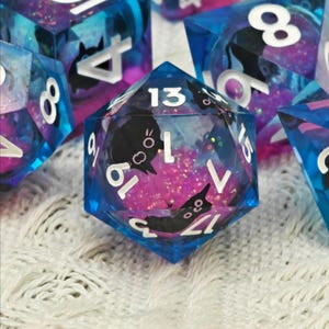 Könnte beinhalten: Satz von Würfeln mit Katzenmotiv. Die Würfel sind transparent mit blauen und rosa Farbtönen, Glitzer und schwarzen Katzensilhouetten. Weiße Zahlen sind auf jeder Seite sichtbar. Ideal für Tischspiele.
