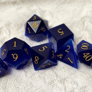 May include: A set of seven polyhedral dice in various shapes, including a d20, d12, d10, d8, d6, and d4. The dice are a deep blue with gold numbering. The dice are arranged on a white, textured surface.