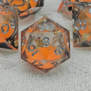 May include: A set of clear resin dice with orange glitter and gold leaf inclusions. The numbers are silver. The dice are for tabletop games.