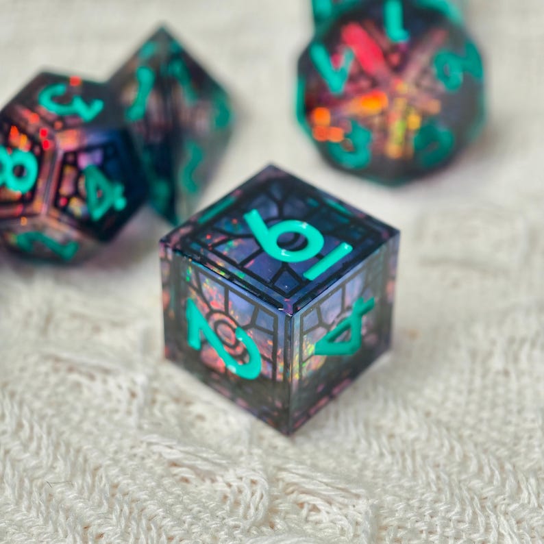 May include: A set of polyhedral dice featuring a dark, translucent resin base with embedded iridescent flakes and bold, bright teal numbers. The cube-shaped die in the foreground displays a black grid pattern resembling stained glass.