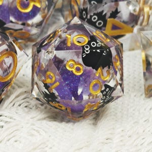 Könnte beinhalten: Ein Satz polyedrischer Würfel mit einem transparenten Harzaußen, goldenen Zahlen und einem lila Glitzerinnenraum. Schwarze Katzengrafiken sind in das Design integriert. Die Würfel sind für Tischspiele konzipiert.