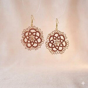Könnte beinhalten: Ein Paar kreisförmige Ohrringe aus braunem, kunstvoll geknotetem Garn in einem floralen Muster. Jeder Ohrring hängt an einem goldfarbenen Metallhaken und zeigt ein filigranes, spitzenartiges Design mit vielen kleinen, symmetrischen Schlaufen.