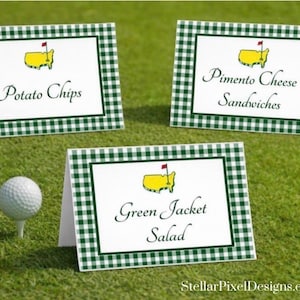 May include: Three white place cards with green gingham borders and the Masters Tournament logo. The cards display the text "Potato Chips", "Pimento Cheese Sandwiches", and "Green Jacket Salad". A golf ball sits on a tee.