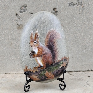 May include: A smooth, oval-shaped gray stone featuring a detailed hand-painted image of a reddish-brown squirrel holding a small pinecone. The squirrel sits on a painted log with fallen leaves, displayed on a black metal wire stand.
