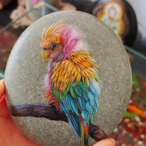 Op de afbeelding: Een handgeschilderde, kleurrijke vogel op een gladde, grijze steen. De vogel heeft een roze kuif, blauwe ogen en levendige gele, groene en blauwe veren. Hij zit op een bruine tak, wat gedetailleerde kunst laat zien.