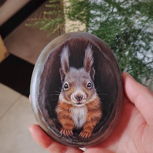 Pode incluir: Uma pedra de rio lisa e oval com uma ilustração detalhada pintada à mão de um esquilo vermelho. O esquilo, com pelo castanho-avermelhado, peito branco e orelhas pontiagudas, é retratado como se estivesse a espreitar de um buraco escuro.