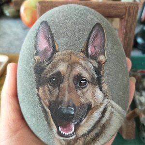 Könnte beinhalten: Ein handgemaltes Porträt eines Deutschen Schäferhundes auf einem glatten, ovalen Stein. Das Gesicht des Hundes ist detailliert mit braunem, schwarzem und tanfarbenem Fell, rosa gefütterten Ohren und einem lächelnden Ausdruck. Der Stein wird in einer Hand gehalten, mit unscharfem Hintergrund.