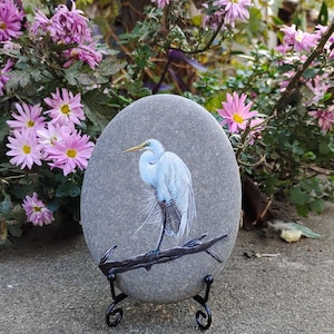 Könnte beinhalten: Ein handbemalter, ovaler grauer Stein zeigt eine detaillierte Darstellung eines weißen Reihers, der auf einem dunklen Ast sitzt. Der Vogel hat einen gelben Schnabel und schwarze Beine. Der Stein wird auf einem schwarzen Metallständer mit rosa Blumen im Hintergrund ausgestellt.