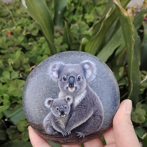Könnte beinhalten: Ein handbemalter, runder grauer Stein zeigt eine Koala-Mutter und ihr Baby. Die Koalas sind in Grau- und Weißtönen dargestellt, mit detailliertem Fell und ausdrucksstarken Augen. Das Kunstwerk ist vor einem verschwommenen grünen Hintergrund.